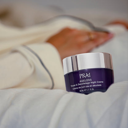 PRAI Beauty Ageless Throat & Decolletage Night Creme with Retinol, jar on a bedsheet beside a resting hand