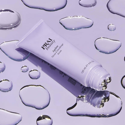 PRAI Beauty Ageless Throat & Decolletage Serum, tube with roller tip on a lilac surface with water droplets