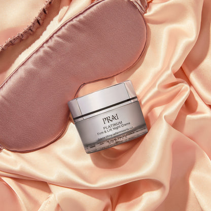 PRAI Beauty Platinum Firm & Lift Night Creme, jar on pink satin beside a sleep mask