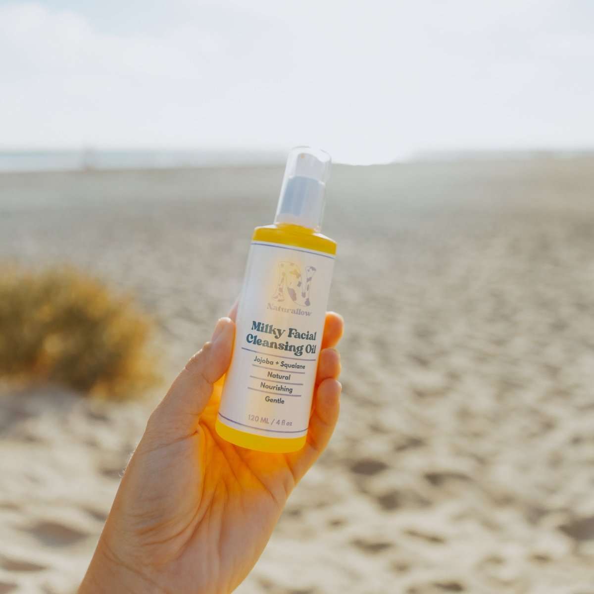 Naturallow Milky Facial Cleansing Oil, hand holding the bottle on the beach