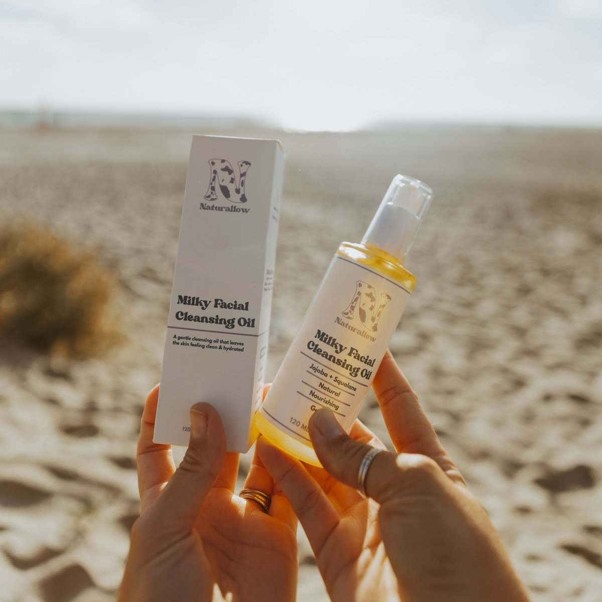 Naturallow Milky Facial Cleansing Oil, hands holding the bottle and box on the beach